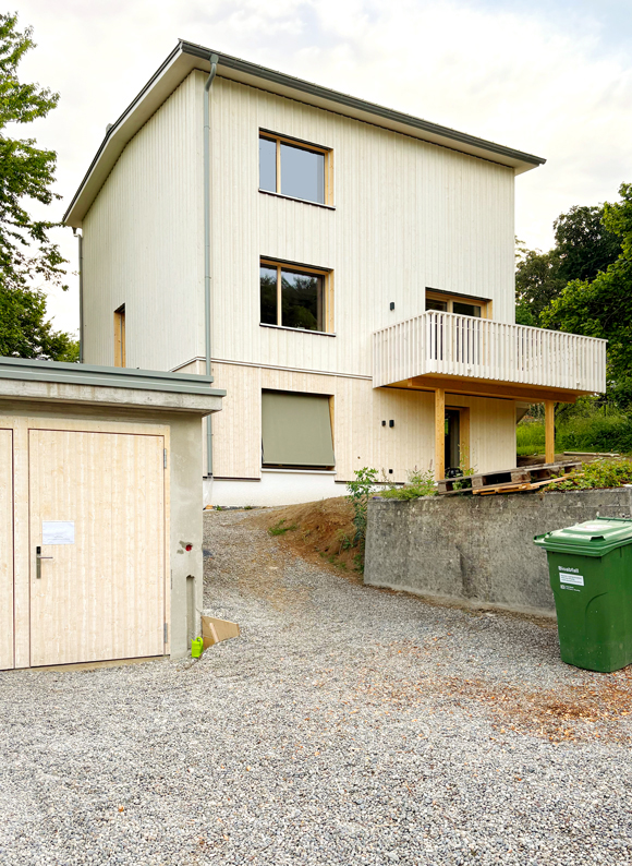 Das freistehende Wohnhaus mit freistehender Garage. Das Wohnhaus mit der neuen Gebäudehülle aus sägerohen Schalungsbrettern weiss geschlämmt mit Schwedenfarbe. Neuer Holzbalkon und erweiterte Garage für Velos. nijo architekten Zürich