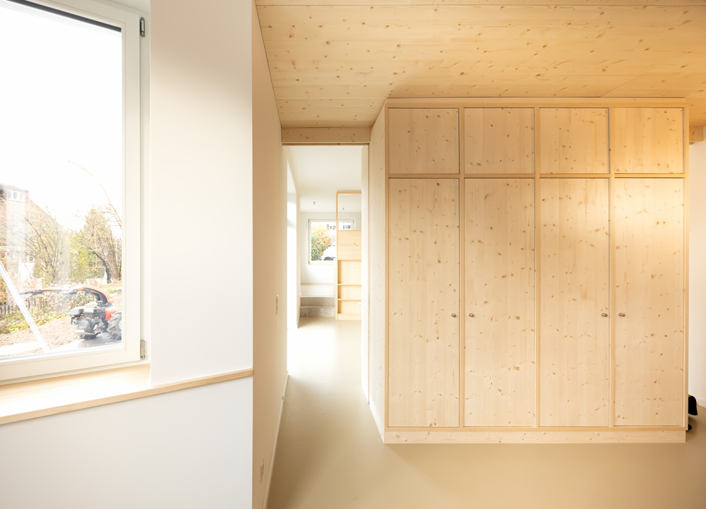 Küsnacht Ausbau Sockelwohnung der offene Raum erstreckt sich über die gesamte Hauslänge, die zwei Treppenstufen aus Sichtbeton ergaben sich aus der Tieferlegung der Bodenplatte, damit mehr Raumhöhe geschaffen werden kann. Gut sichtbar die beiden Raumteiler Holzküchenzeile und Holzkleiderschrank, welche beidseitig passierbar sind. Im Bereich des Holzschrankes war ein Stahlträger notwendig um den Übergang von Hourdisdecke zu Betondecke aufzufangen. Darunter laufen neu die beidseitigen Schiebetüren.